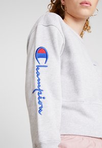 Sweatshirt gris avec un logo "Champion" bleu brodé et un emblème circulaire coloré sur la manche. Tissu texturé avec une coupe décontractée.