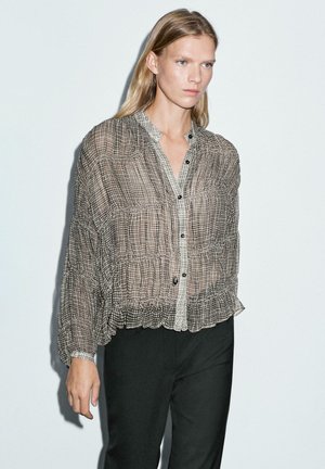 Blonde vrouw in een doorzichtige, geblokte blouse met knopen en opgeblazen mouwen, in een zwarte broek, staande tegen een witte achtergrond.