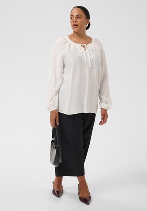 Femme portant un chemisier blanc à manches longues avec des détails en dentelle, un pantalon noir à jambes larges, des chaussures pointues foncées et un sac à main noir.