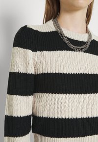 Pull en tricot rayé noir et crème avec une texture côtelée. Il dispose d'un col rond et est associé à un collier en chaîne argentée multi-couches.