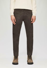 Braune, schmal geschnittene Hosen aus glattem Stoff, mit Fronttaschen und einem Knopfverschluss. Kombiniert mit beige-farbenen Stiefeln für einen Kontrast.