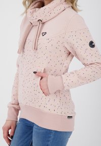 Světle růžová mikina s kapucí a barevným skvrnitým vzorem. Obsahuje přední klokaní kapsu, žebrované manžety a štítek značky na boku.