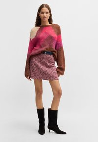 Hrubý pletený sveter v hnedých, ružových a krémových zygzagových vzoroch; skombinovaný s červenou tweed minisukňou; čierne zdobené čižmy po kolená.