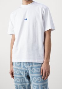 T-shirt bianca in cotone con un piccolo logo blu "HUGO" sul petto, abbinata a jeans di denim blu chiaro con un audace motivo di testo ripetuto.