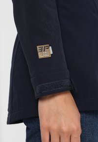 Manche de veste marine en tissu texturé, avec une étiquette beige à lettres noires et une fermeture à scratch sombre au poignet.