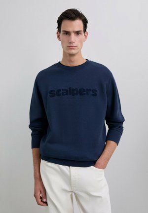 Joven vestido con un sudadero azul marino que lleva el texto "scalpers" y pantalones crema, de pie con una mano en el bolsillo frente a un fondo liso.