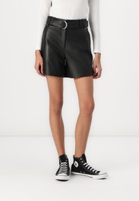 Shorts en cuir noir avec une taille ceinturée présentant une boucle circulaire, associés à des baskets montantes noires avec des semelles blanches et un logo.