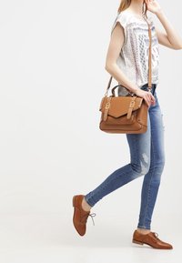 Bruine leren schoudertas met dubbele gespaccenten, crossbody gedragen. Model in licht graphic T-shirt en blauwe versleten jeans, lopend.