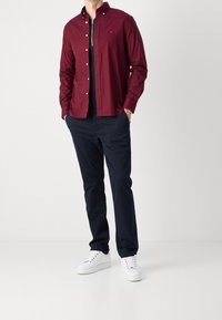 Burgundowa koszula z długim rękawem z guzikami, w połączeniu z granatowymi spodniami i białymi trampkami. Prosty design, gładka faktura i standardowy krój.