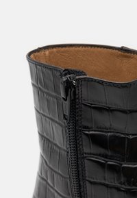 Bota de tobillo de piel negra con textura de cocodrilo, que cuenta con un cierre lateral con cremallera y un forro interior marrón. Acabado suave y diseño elegante.