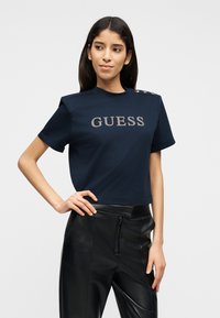 Γυναίκα με μπλε ναυτικό μπλουζάκι κοντομάνικο με το λογότυπο "GUESS" και μαύρο δερμάτινο παντελόνι, πόζα με το ένα χέρι στη μέση μπροστά σε λευκό φόντο.