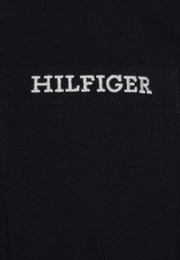 Mustan tekstuurin kangas, jossa valkoisella brodeerattu teksti "HILFIGER" on keskitetty ommellulle suorakaiteen muotoiselle tarralle.