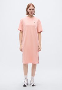 Ροζ βαμβακερό φόρεμα t-shirt με κοντά μανίκια, ελαφρώς ανοίγματα στο πλάι και μικρό λογότυπο στο στήθος. Συνδυάζεται με λευκά αθλητικά παπούτσια και κάλτσες αστραγάλου.