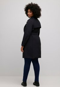 Czarny trencz z paskiem w talii, zapięciem na guziki przy mankietach oraz wentylacją z tyłu. Połączony z niebieskimi dżinsami i czarnymi botkami do kostek.