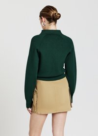 Maglione verde lavorato a maglia con colletto e orlo a coste, abbinato a una mini gonna beige con tasche laterali e una texture liscia.