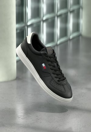 Baskets basses noires avec semelle blanche et logo rouge, blanc et bleu, exposées au-dessus d'un sol gris réfléchissant près de blocs de verre.