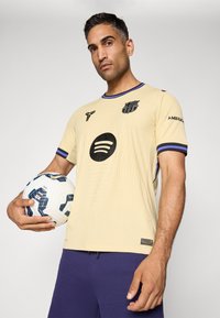 Flødefarvet fodboldtrøje med et tekstureret design, sort Spotify-logo og lilla accenter. Spilleren holder en hvid og blå fodbold.