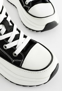 Czarno-białe sneakersy z teksturowanym płóciennym korpusem, masywną białą podeszwą, okrągłym czubkiem i białymi sznurówkami przewleczonymi przez srebrne oczka.