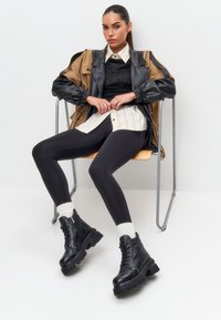 Robuuste zwarte leren enkellaarzen met een vetersluiting en dikke zolen, gedragen met zwarte leggings en een oversized jas in bruin en zwart.