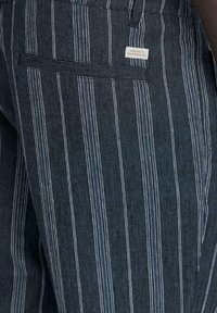 Pantalon en denim foncé avec des fines rayures verticales blanches et bleues, et une poche arrière étiquetée "Dédié à la perfection du denim."