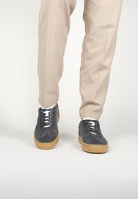 Zapatillas de ante en gris oscuro con suela de goma color caramelo, con cordones grises y una lengua blanca, combinadas con pantalones beige claros.