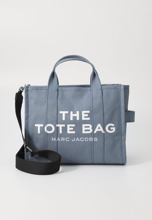 Geantă de tote din pânză albastră, cu două mânere superioare, curea neagră detașabilă și branding alb "THE TOTE BAG", având un design structurat și cusături.