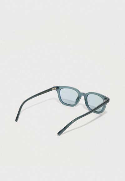 Lunettes de soleil à monture carrée translucide bleue avec des verres bleu clair reposant ouvertes sur une surface blanche.