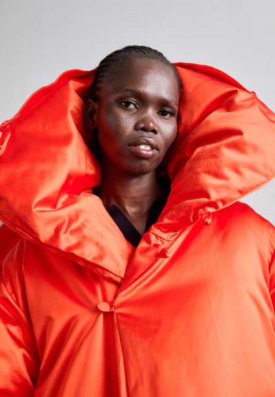 Λαμπερό πορτοκαλί μπουφάν με πλούσιο γιακά, φτιαγμένο από γυαλιστερό ύφασμα. Διαθέτει κλείσιμο με κουμπί και υφή στην επιφάνειά του.