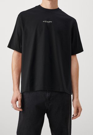 Personne portant un t-shirt noir G-Star Raw avec un petit logo blanc sur la poitrine et un pantalon noir sur un fond gris.