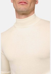 Pull en col roulé crème en tissu doux et côtelé. Design ajusté avec un col haut, mettant en avant une texture lisse et une apparence minimaliste.