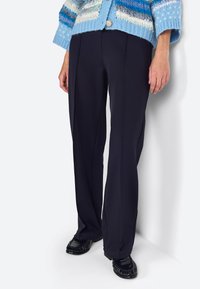 Pantaloni navy su misura con taglio dritto, caratterizzati da una texture liscia e delicate pieghe frontali, abbinati a scarpe nere in vernice.