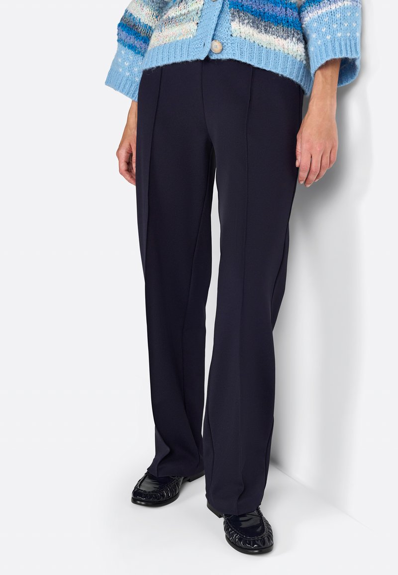 Pantaloni navy su misura con taglio dritto, caratterizzati da una texture liscia e delicate pieghe frontali, abbinati a scarpe nere in vernice.