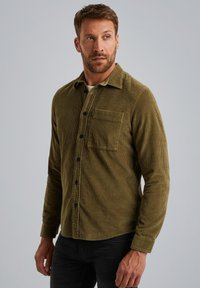 Groene corduroy shirt met een kraag, voorkant pocket, zwarte knopen en gestructureerde stof. Lange mouwen met knoopmanchetten. Model draagt een zwarte broek.