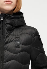 Κοντινό πλάνο ατόμου που φοράει μαύρο καπιτονέ μπουφάν με κουκούλα, εμπρόσθιο φερμουάρ και μικρή ετικέτα «Blauer U.S.A.» στο στήθος.