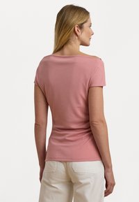 Top aderente a maniche corte in morbido tessuto rosa, con scollo ampio e design drappeggiato sul retro, abbinato a pantaloni chiari.