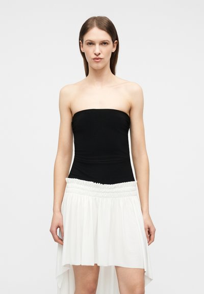 Γυναίκα που φοράει στενό μαύρο τοπάκι χωρίς τιράντες σε συνδυασμό με λευκή φούστα με πιο μακρύ πίσω μέρος, στέκεται μπροστά σε απλό ανοιχτό γκρι φόντο.