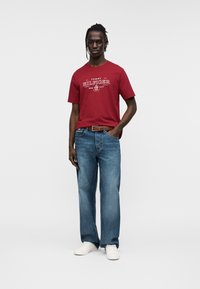 Κόκκινο βαμβακερό t-shirt με λευκό τυπωμένο λογότυπο, συνδυασμένο με μπλε τζιν παντελόνι ευθείας γραμμής και λευκά αθλητικά παπούτσια. Το μοντέλο στέκεται με τα χέρια στις τσέπες.