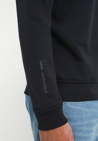 Svart hettegenser med ribbestruktur, med en synlig Karl Lagerfeld-logo på venstre erme, paret med blå denimjeans.