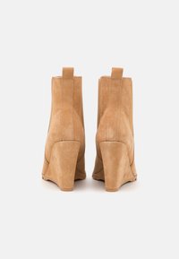 Botas de tornozelo em cunha feitas em camurça bege, com um salto curvo, alça traseira e costura vertical para adicionar textura e detalhe.