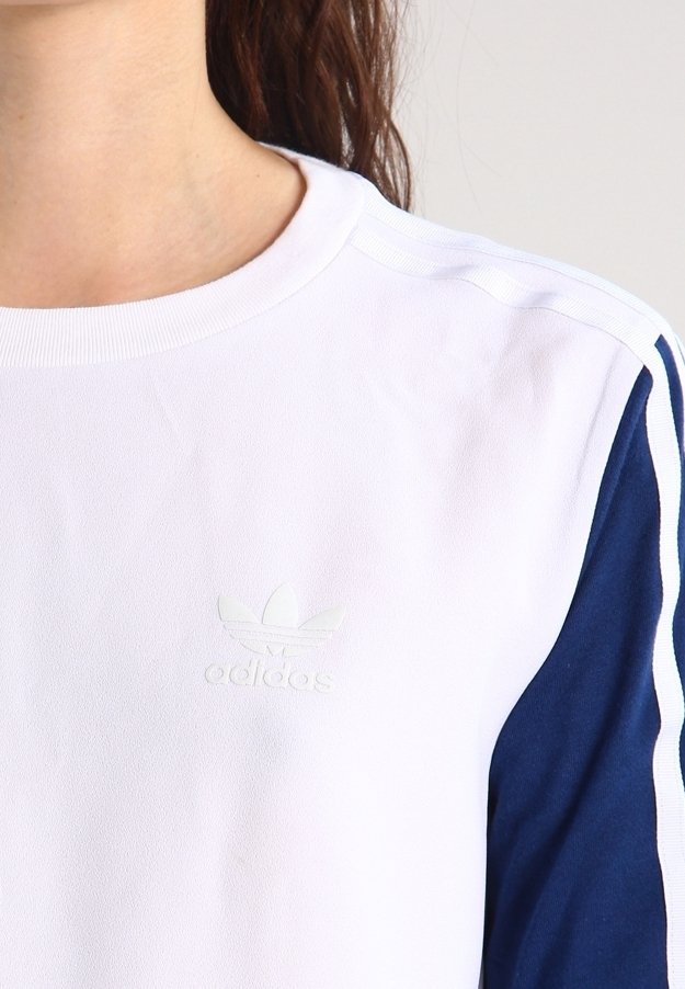 Vit sweatshirt med marinblå ärmar, ribbad krage och en strukturerad Adidas-logotyp på bröstet. Har kontrasterande sidostrippar.