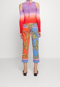 Rippenshirt mit kaltem Schulterdesign, das von Lila nach Rot verläuft. Bunte, gemusterte Jeans mit orangefarbenen und blauen Abschnitten, goldenen Akzenten und tailliertem Schnitt.