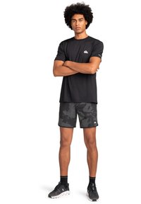 Černé sportovní tričko s krátkým rukávem, doplněné o tmav�é kraťasy s maskáčovým vzorem. Model má na sobě černé tenisky a stojí se založenýma rukama.