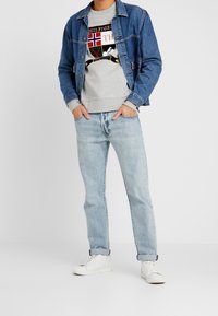 Chaqueta de denim sobre sudadera gris con logo, jeans de mezclilla claros y zapatillas blancas. Atuendo casual con un corte relajado y texturas suaves.