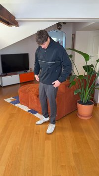 Svart genserjakke med glidelås og refleksstriper, grå rutete bukser og hvite joggesko; innendørs med en grønn plante og oransje sofa.