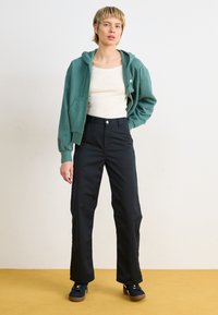 Green zip-up hoodie over a white tank top, paired with high-waisted black pants. Black sneakers with a brown rubber sole.