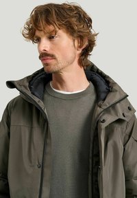 Olivgrön jacka med dragkedja fram och huva, med svart foder och knappar. Bärs över en texturerad grå tröja.