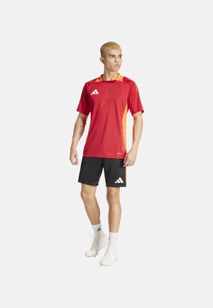 Rode sporttrui met oranje accenten, zwarte shorts en witte voetbalschoenen. Bevat het Adidas-logo op de borst en de shorts, met korte mouwen en een ronde hals.