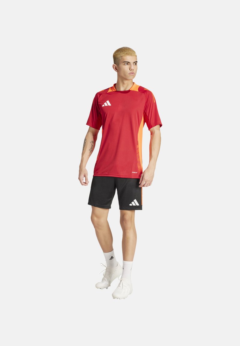 Rotes Sporttrikot mit orangefarbenen Akzenten, schwarze Shorts und weiße Fußballschuhe. Mit Adidas-Logo auf der Brust und Shorts, kurzen Ärmeln und Rundhalsausschnitt.