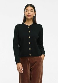 Cardigan noir tricoté avec un col rond, des boutons dorés sur le devant et des manches longues. Associé à un pantalon en velours côtelé marron.