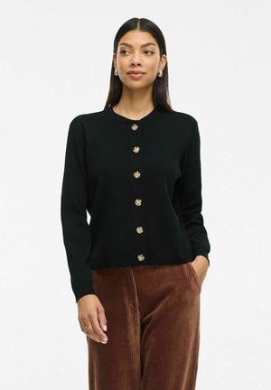 Black knitted cardigan with a round neckline, gold buttons down the front, and long sleeves. Paired with brown corduroy trousers.